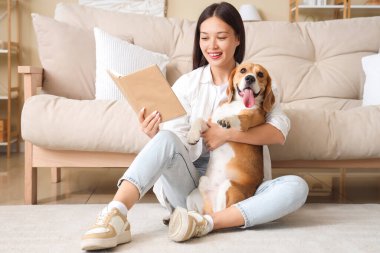 Kitaplı genç bir kadın oturma odasında yerde sevimli bir Beagle köpeğine sarılıyor.