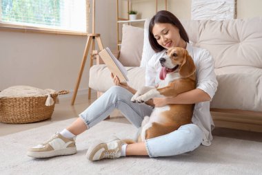 Kitaplı genç bir kadın oturma odasında yerde sevimli bir Beagle köpeğine sarılıyor.