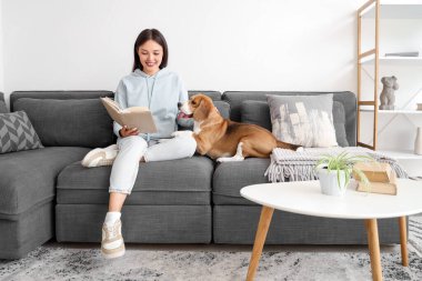 Genç bir kadın kitap okuyor ve oturma odasındaki kanepede sevimli bir tazı köpeğiyle oturuyor.