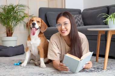 Gözlüklü genç kadın kitap okuyor ve oturma odasında yerde tapılası Beagle köpeğiyle yatıyor.