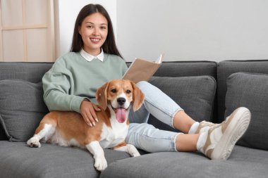 Genç bir kadın kitap okuyor ve oturma odasındaki kanepede tatlı bir tazı köpeğiyle dinleniyor.