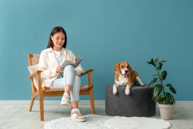 Oturma odasında, sevimli Beagle köpeğinin yanındaki koltukta kitap okuyan genç bir kadın.