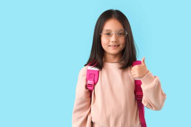 Sırt çantalı şirin mutlu Asyalı kız mavi arka planda baş parmak hareketi gösteriyor. Okul yılı sonu.