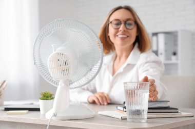 Ofisinde elektrikli yelpaze ve bir bardak su olan güzel, olgun bir iş kadını.