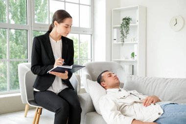 Ofiste hipnotize edilmiş bir hastayla deftere yazan kadın psikolog.