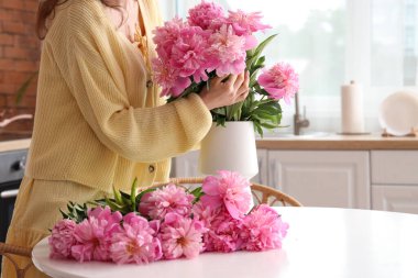 Genç kadın şakayık demetini mutfaktaki masanın üzerine vazoya koyuyor.