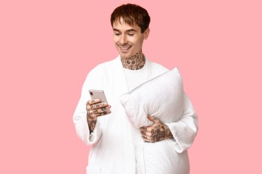 Pembe arka planda yastıklı ve cep telefonlu pijamalı dövmeli genç adam.