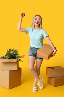 Sarı arka planda karton kutuları ve anahtarları olan mutlu genç bir kadın. Yeni ev konseptine geçiyoruz.