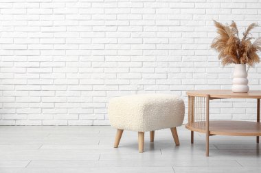 Beyaz tuğla duvarın yanında vazo ve puf kaplı sehpa.