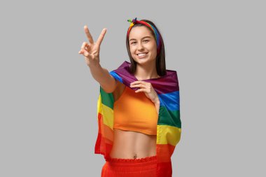 Göbeğinde piercing ve LGBT bayrağı olan genç ve güzel bir kadın gri arka planda zafer jesti gösteriyor.