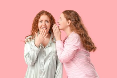 Pembe arka planda dedikodu yapan pijamalı mutlu kızıl kız kardeşler.