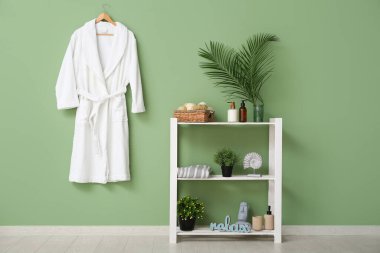 Yeşil duvarda asılı beyaz bornoz ve banyoda raflarda farklı banyo malzemeleri.