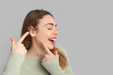 Gri arka planda işitme cihazına işaret eden mutlu genç kadın