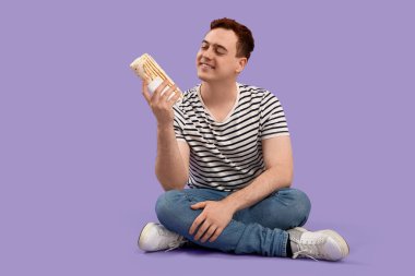 Young man sitting and holding doner kebab on purple background