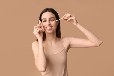 Beautiful young woman applying concealer on brown background
