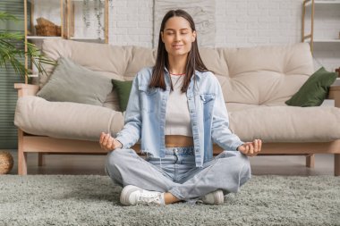 Genç bir kadın oturma odasındaki kanepenin yanında meditasyon yapıyor.