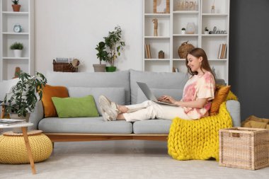 Genç bir kadın oturma odasındaki gri koltukta dizüstü bilgisayar kullanıyor.