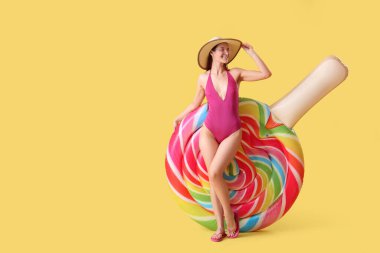 Smiling woman in bikini and hat with inflatable mattress on yellow background