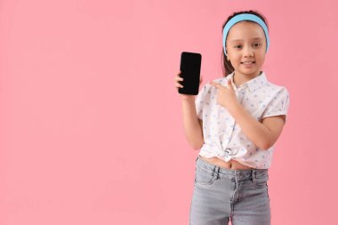 Pembe arka planda boş ekranlı cep telefonuna bakan zarif küçük bir kızın portresi.