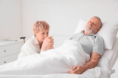 Klinikte hasta kocasının elini tutan olgun bir kadın.