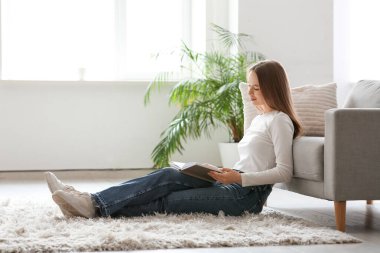 Oturma odasındaki kanepenin yanında kitap okuyan genç bir kadın.