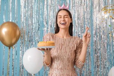 Doğum günü pastası ve balonlarıyla mutlu genç bir kadın mavi arka planda dilek diliyor.