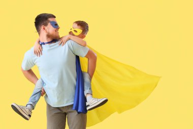 Sarı arka planda süper kahraman kostümlü babasıyla küçük bir çocuk.