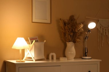 Vazonun yanında parlayan lamba. Odadaki çekmecelerin üzerinde otlar var. Yakın plan.