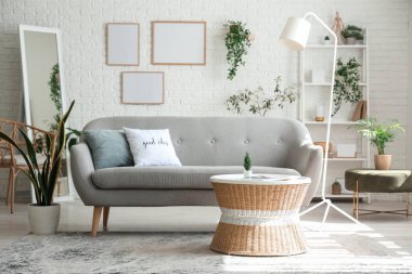 Aydınlık oturma odasının içinde gri kanepe, sehpa ve beyaz tuğla duvarda boş çerçeveler.