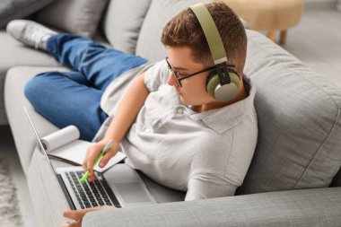 Evde çalışan dizüstü bilgisayarı ve kulaklığı olan genç bir çocuk.