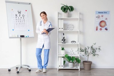 Klinikte göz testi tablosunun yanında panosu olan kadın göz doktoru.