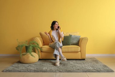 Güzel genç bir kadın cep telefonuyla konuşuyor ve evdeki sarı kanepede dinleniyor.