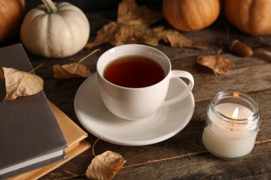 Kitaplı bir fincan çay, sonbahar yaprakları ve ahşap arka planda yanan bir mum.