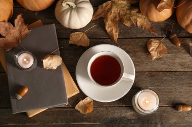Bir fincan çay, kitaplar, sonbahar yaprakları, yanan mumlar ve ahşap arka planda balkabakları.