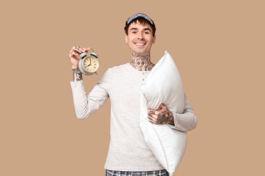 Kahverengi arka planda yastıklı ve çalar saati olan pijamalı dövmeli genç bir adam.