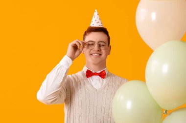 Parti şapkalı, balonlu mutlu genç adam sarı arka planda doğum gününü kutluyor.