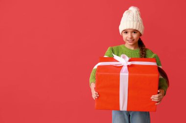 Kırmızı arka planda Noel hediyesi olan sıcak şapkalı şirin küçük kız.