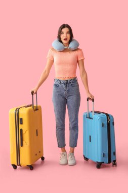 Güzel, genç, şok olmuş bir bayan turist bavulları ve pembe arka planda seyahat yastığı.