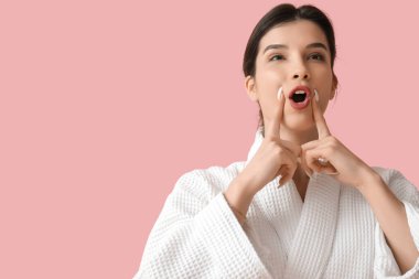 Bornozlu güzel bir kadın pembe arka planda yüz geliştirme egzersizi yapıyor.