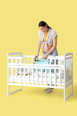 Sarı arka planda bebek beşiğinde doğum çantası taşıyan hamile bir kadın.
