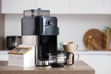 Mutfaktaki kahve makinesinde fincanlar, şeker ve bir kavanoz fasulye var.