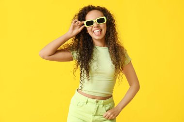 Güneş gözlüklü genç Afro-Amerikalı kadın sarı arka planda dilini gösteriyor.