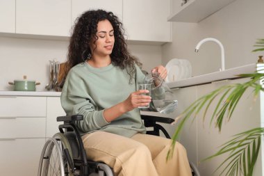 Tekerlekli sandalyedeki genç Afro-Amerikalı kadın evdeki sürahiden cama su dolduruyor.