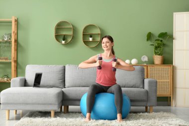 Oturma odasında halterli güzel bir genç bayan antrenman yapıyor.