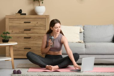 Dizüstü bilgisayarlı güzel bir kadın oturma odasında yoga yapıyor.