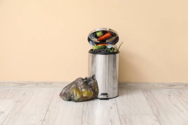 Gıda atıkları ve bej duvarın yanında dolu çöp torbası olan metal çöp kutusu. Geri dönüşüm kavramı