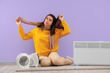 Güzel, terli, kışlık elbiseli, radyatörü ve elektrikli fan ısıtıcısı olan bir kadın mor duvarın yanında oturuyor.