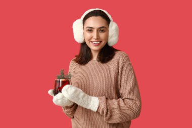 Kırmızı arka planda sıcak sıcak şarap kadehiyle sıcak eldivenler içinde mutlu genç bir kadın.