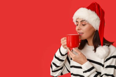 Kırmızı arka planda sıcak şarap bardağıyla Noel Baba şapkalı genç bir kadın.