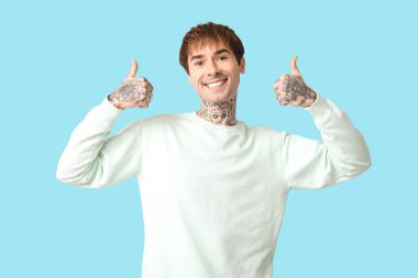 Mutlu, yakışıklı, dövmeli adam mavi arka planda baş parmak işareti yapıyor.